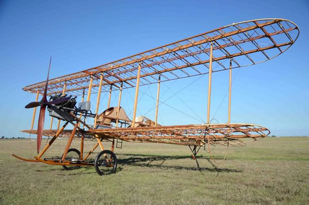 A wooden-framed replica of the Royal Aircraft Factory BE2a biplane, its wings without fabric covering, on an open grass airfield.